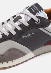 Grauer und beige Sneaker aus Mesh- und Wildledermaterialien. Verfügt über graue Schnürsenkel, ein Logo an der Seite und eine strukturierte Gummisohle.