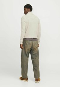 Suéter de punto color crema con cuello alto, usado sobre una camiseta oscura, combinado con pantalones de mezclilla verde desgastados y zapatos casuales marrones.