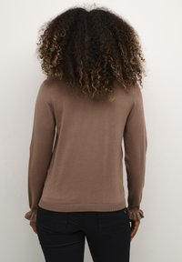 Ruskea pitkähihaisessa villapaidassa on ribbikuvioiset hihansuut ja helma, se on sileä ja tiukasti istuva malli, yhdistettynä tummiin housuihin.