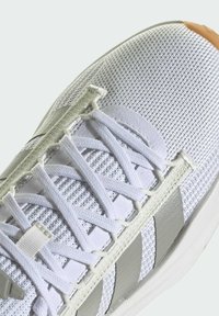 Zapatilla deportiva blanca de malla con rayas grises, cordones planos y acabado texturizado. La puntera presenta una tapa de goma en color marrón claro para mayor durabilidad.
