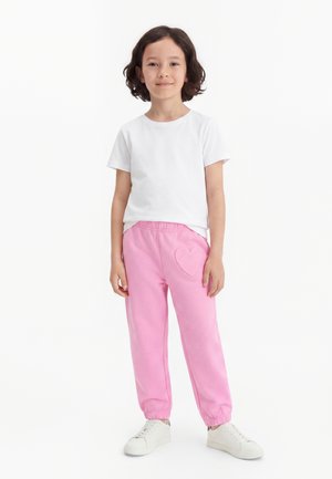 Kind mit dunklen Haaren, das ein weißes T-Shirt, rosa Hose mit Herz-Aufnäher und weiße Sneakers trägt, steht vor einem schlichten weißen Hintergrund.