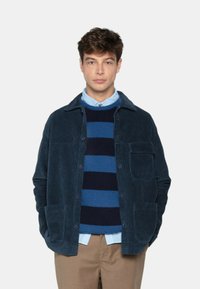 Jeune homme portant une veste en velours côtelé bleu, un pull rayé bleu et marine, une chemise bleu clair et un pantalon beige, se tenant avec les mains dans les poches.