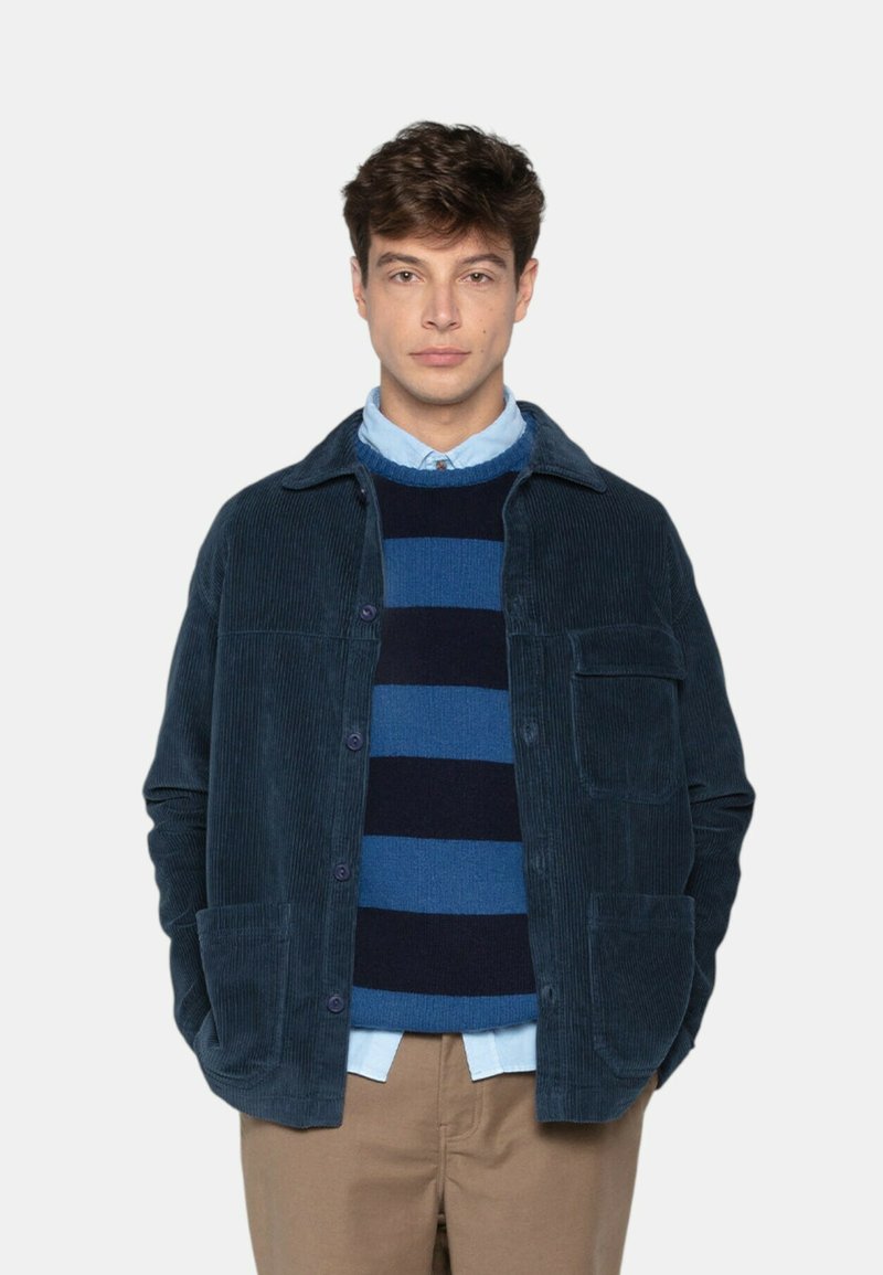 Jeune homme portant une veste en velours côtelé bleu, un pull rayé bleu et marine, une chemise bleu clair et un pantalon beige, se tenant avec les mains dans les poches.