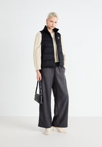 Gilet noir sur un pull en maille crème, associé à un pantalon large gris et des bottines beiges à talons, portant un petit sac à main noir.