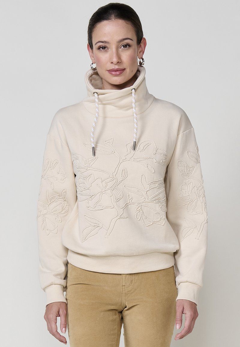 Sweatshirt beige avec col montant, orné de broderies florales sur le devant et les manches. Agrémenté de cordons de serrage et de poignets côtelés.