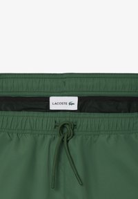 Shorts de bain verts avec taille élastique, fermeture à cordon, et doublure en maille noire. Présente une étiquette logo Lacoste à la taille.