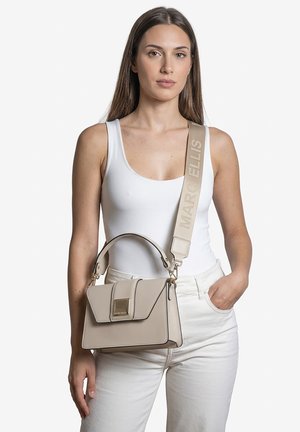 Femme portant un débardeur blanc et un pantalon, portant un sac à main beige Marc Ellis avec une large bandoulière de marque sur l'épaule.