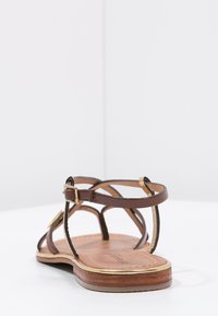Sandalo in pelle marrone con cinturini, suola piatta, dettagli dorati e chiusura con fibbia. Presenta una texture liscia e un design aperto.
