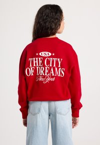 Giovane con lunghi capelli scuri che indossa una felpa rossa con scritto bianco "USA LA CITTÀ DEI SOGNI New York" e jeans blu chiaro, in piedi.