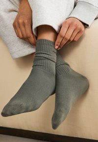 Mains ajustant des chaussettes grises côtelées sur des pieds, personne portant un pantalon de survêtement gris clair, assise sur un canapé beige.