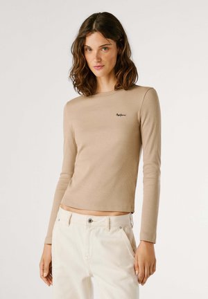 T-shirt beige à manches longues en tissu côtelé avec un col rond et une coupe courte. Présente un petit logo noir sur la poitrine. Associé à un pantalon blanc.