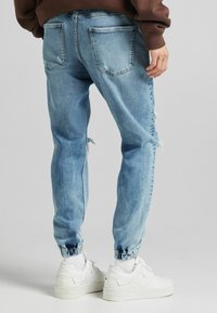 Person som bär ljusblå slitna denim-joggingbyxor med elastiska ärmar, vita sneakers och en brun sweatshirt.