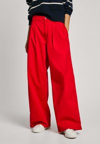 Pantaloni larghi rossi con pieghe, realizzati in tessuto morbido. Abbinati a un maglione a righe blu navy e scarpe bianche. Presentano una chiusura con bottone.