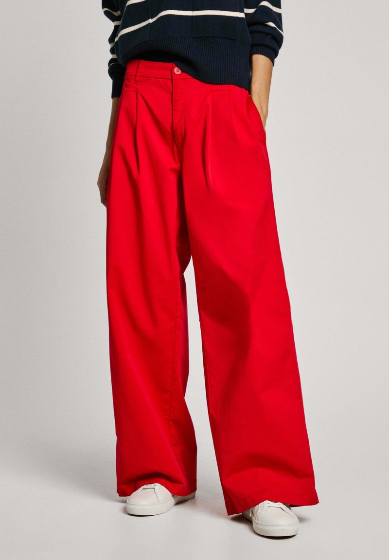 Pantaloni larghi rossi con pieghe, realizzati in tessuto morbido. Abbinati a un maglione a righe blu navy e scarpe bianche. Presentano una chiusura con bottone.