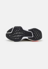 Semelle de chaussure de sport noire et blanche présentant une surface en caoutchouc texturé avec des motifs variés et un accent rouge au talon.