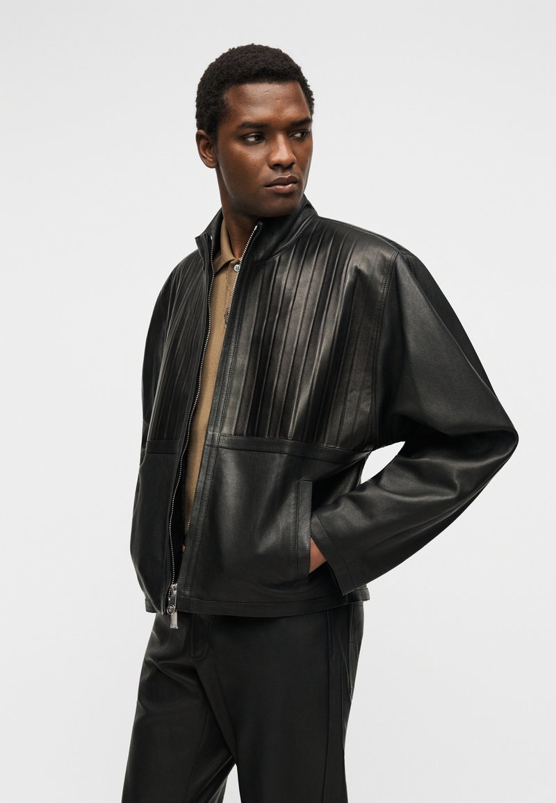 Homme portant une veste en cuir noire avec un matelassage vertical sur la poitrine et un pantalon en cuir noir assorti, regardant sur le côté.