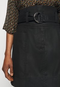 En svart kjol i texturerat tyg, med en bred midjeband och en silverfärgad rund spänne samt sidofickor, stajlad med en blommig blus.