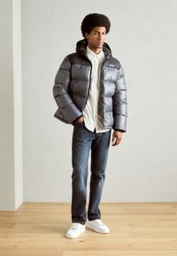Grå pufferjakke med glidelås og hette, kombinert med en hvit skjorte med knapper, blå jeans og hvite joggesko på et tregulv.