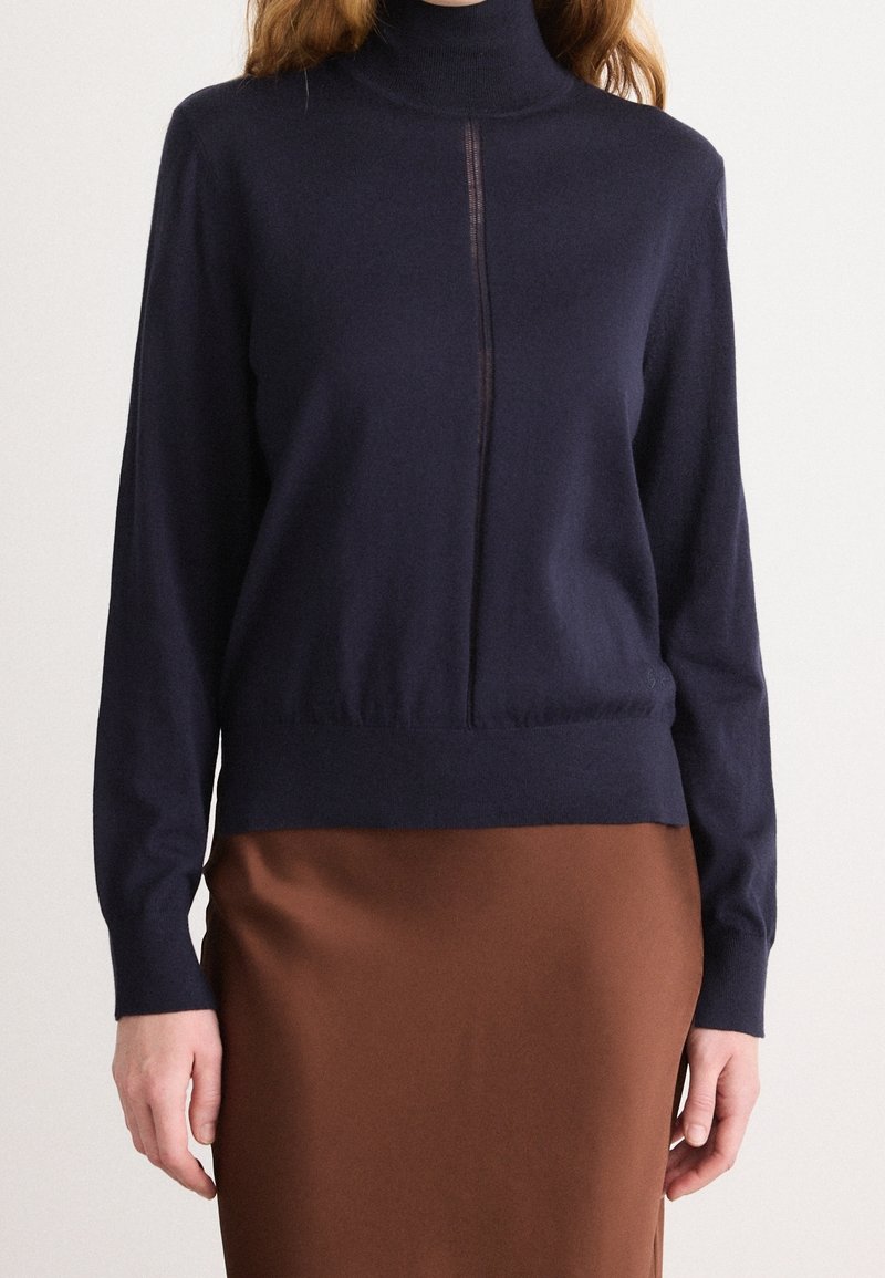Femme portant un pull à col roulé bleu marine avec une couture centrale, associé à une jupe marron, debout devant un fond uni.