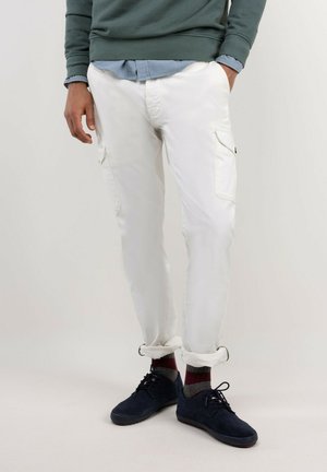 Hombre con pantalones cargo blancos con puños remangados, calcetines oscuros, zapatos azul marino y sudadera verde sobre una camisa azul.
