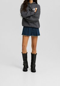 Sudadera gris con capucha y un gráfico, combinada con una falda de mezclilla con pliegues y botas negras hasta la rodilla con correas decorativas y tachuelas.
