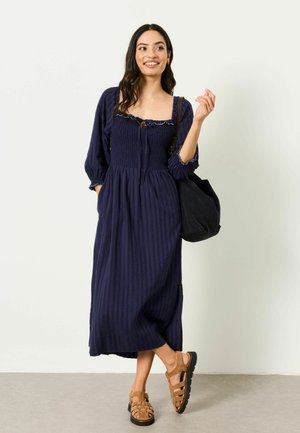 Frau in einem marineblauen Midikleid mit 3/4-Ärmeln, die eine schwarze Umhängetasche trägt und beige Sandalen anhat, steht vor einer weißen Wand.