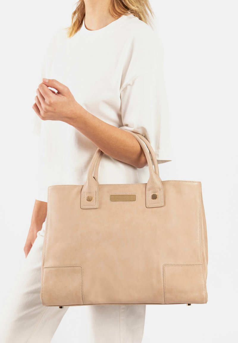 Sac tote en cuir beige avec deux poignées supérieures, forme carrée, détail de poche frontale et accents de couture subtils. Texture lisse et design minimaliste.