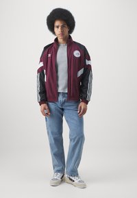 Corta-vento marrom e preto com riscas brancas, logótipo prateado, usado sobre uma camisola cinza de nervuras, combinado com jeans azul claro e sapatilhas.