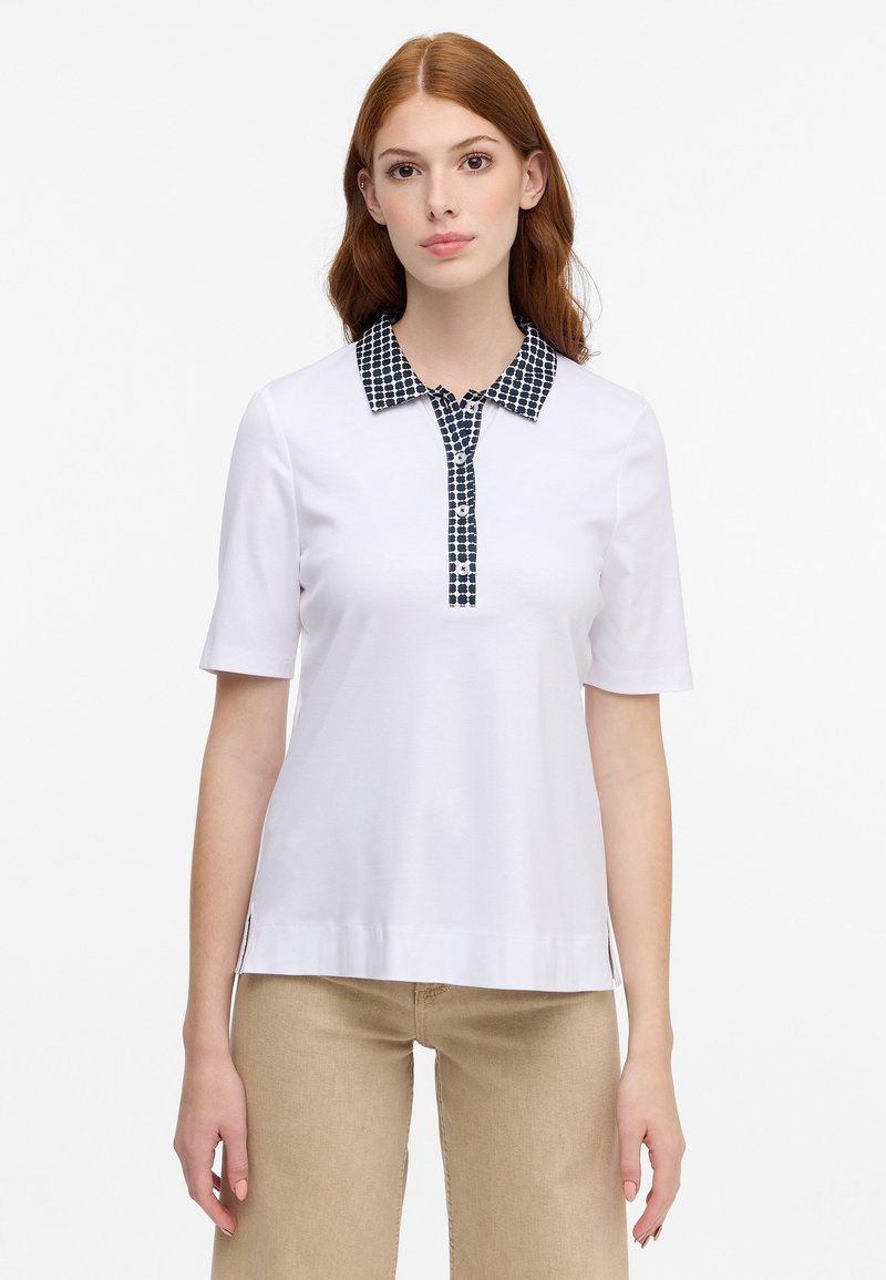Maglietta polo bianca con colletto a quadri blu e placca con tre bottoni. Maniche corte e orlo dritto con spacchetti laterali per facilitare i movimenti.