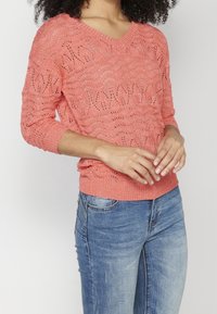 Pull en maille corail avec un col en V et des manches trois-quarts, présentant un motif texturé. Associé à un jean en denim bleu.