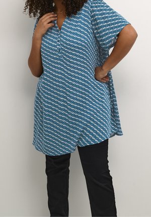 Turquoise tuniek met een wit kraanvogelpatroon, V-hals, korte mouwen en een losse pasvorm; gedragen met zwarte strakke broeken.