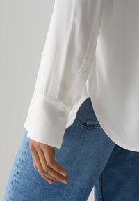 Weißes Langarmhemd mit einer geknöpften Manschette, weicher Textur und leicht abgerundetem Saum, kombiniert mit blauen Jeans.