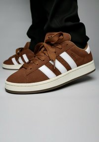 Brown suede sneakers with white stripes and laces, featuring a textured upper and a light-colored rubber sole. Brand label visible.