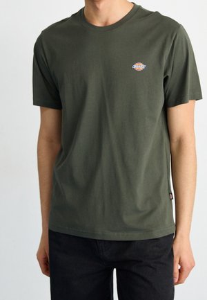 Camiseta de algodón verde oliva con mangas cortas, escote redondo y un pequeño parche de logo en el lado izquierdo del pecho. Combinada con pantalones negros.