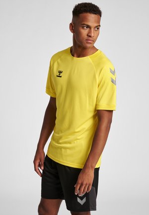 Giovane uomo con maglietta sportiva gialla e pantaloncini neri, che guarda di lato su uno sfondo chiaro uniforme.