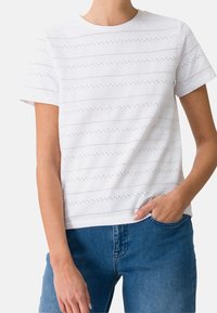 T-shirt blanc à manches courtes avec des motifs à découpe horizontale et un col rond, associé à un jean en denim bleu.