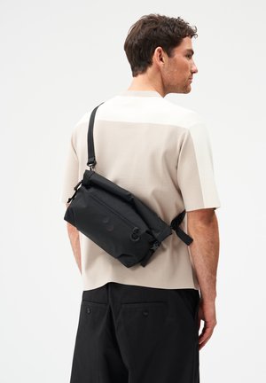 Mann mit beigefarbenem Kurzarmhemd und schwarzer Hose, der eine schwarze Umhängetasche auf dem Rücken trägt, seitlich vor einem einfarbigen Hintergrund stehend.