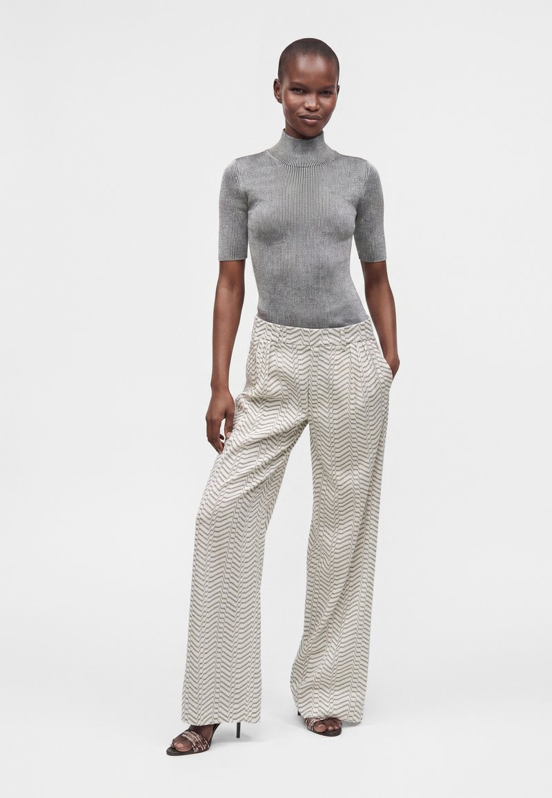 Femme portant un col roulé gris ajusté à manches courtes et un pantalon ample à motifs, debout avec une main dans la poche et des sandales à talons hauts.