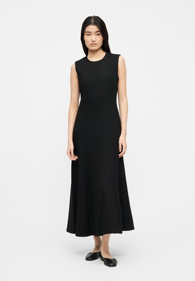 Naine seisab sirgete pikkade mustade juustega, kandes varrukateta musta midi-pikka kleiti ja musti madalaid kingi tavalise valge tausta ees.