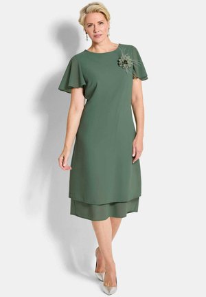 Frau trägt ein knielanges grünes Kleid mit schwingenden Ärmeln und einer Blumenbrosche auf der Brust, kombiniert mit silbernen High Heels.