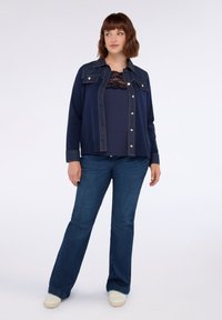 Donna in piedi con indosso un giubbotto blu scuro a bottoni sopra una maglietta navy con pizzo, jeans blu e sneakers bianche su uno sfondo bianco.