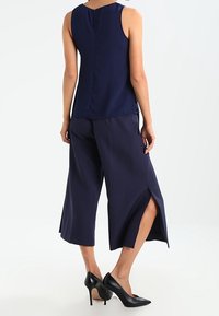 Marinblå utan ärm topp i kombination med vida marinblå byxor med sidslitsar, bärs med svarta högklackade skor. Slät tyg, minimalistisk design.