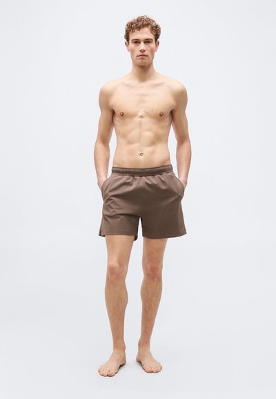 Νεαρός άνδρας που στέκεται ξυπόλυτος μπροστά σε απλό φόντο, φορώντας καφέ σορτς με ελαστική μέση και έχοντας τα χέρια στις τσέπες.