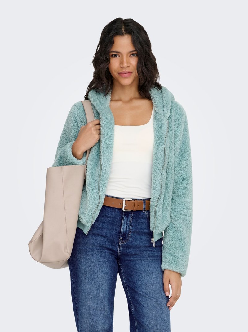 Femme portant une veste à capuche douce de couleur teal, un haut blanc, un jean bleu et une ceinture marron, tenant un grand sac fourre-tout beige sur son épaule.
