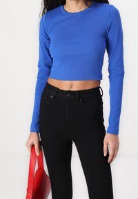 Top corto a maniche lunghe blu a coste abbinato a jeans neri a vita alta, accompagnato da una borsa rossa. Il materiale sembra elastico, con un design aderente.