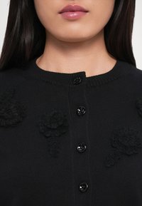 Mustat neulotut kardiganit, joissa on kolmiulotteisia kukka-applikaatioita, pyöreä kaula-aukko ja mustat napit edessä.