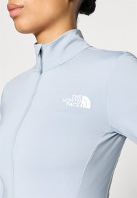 Ανοιχτό μπλε αμάνικο κορμάκι απόδοσης με μακριά μανίκια, ψηλό γιακά και ημι-φερμουάρ κλείσι μο. Διαθέτει λευκό λογότυπο στην αριστερή περιοχή του στήθους.