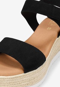Sandalia de ante negra con dos correas anchas, una plantilla color tostado y una suela de plataforma estilo yute tejido. La suela cuenta con un fondo de goma blanca.
