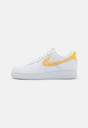 Hvid lave sneakers med gul børstet logo og gul hæltab, vist fra ydersiden på en hvid baggrund.