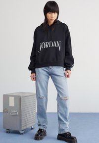 Sudadera negra con el texto "JORDAN" en blanco, bolsillo delantero, combinada con jeans desgastados de color azul claro y zapatos de plataforma negros. Maleta plateada cercana.